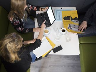 Zwei Frauen und ein Mann sitzen am Tisch und arbeiten mit Laptop und Broschüren.