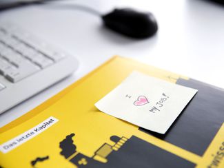  Schreibtisch mit  Tastatur, Computer-Maus und Broschüre, darauf ein Zettel mit der Inschrift "I love my job!"